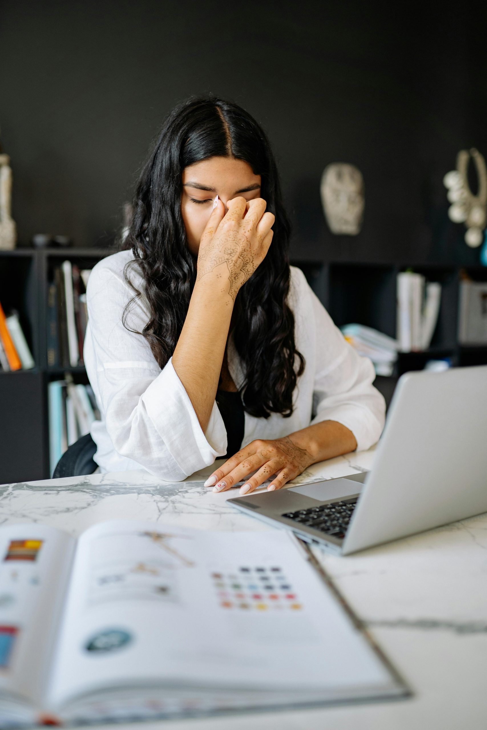 A woman feeling stressed while working on a laptop in an office setting with documents.
